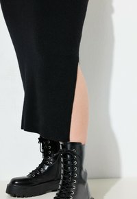 Benen in zwarte mid-calf gevechtslaarzen, gecombineerd met een zwarte rok met een zijspleet, tegen een witte muur als achtergrond.
