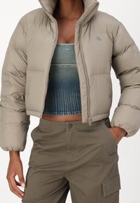 Giacca puffer beige con colletto alto e chiusura a zip, con un dettaglio del logo. Sotto, un top blu corto con una texture a coste. Pantaloni cargo olive.