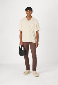 Beige Poloshirt mit kurzen Ärmeln und Brusttasche, braune Slim-Fit-Hosen und hellfarbene Sneakers. Schwarze kleine Umhängetasche wird in der Hand gehalten.