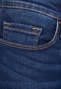 Detalle del bolsillo trasero de unos vaqueros, tela azul oscura, costuras visibles en naranja, textura suave y pequeño bolsillo adicional por encima del principal.