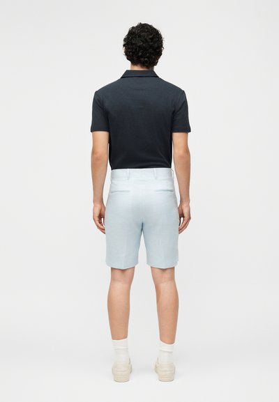 Άνδρας με σγουρά μαλλιά που φοράει μαύρο πόλο με κοντά μανίκια, ανοιχτό μπλε σορτς, λευκές κάλτσες και μπεζ αθλητικά παπούτσια, στέκεται με την πλάτη προς τα εμάς.