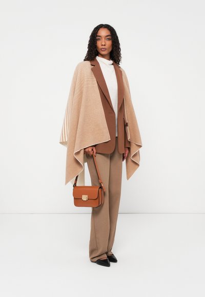 Beige ullponcho med ribbede detaljer, kombinert med en brun blazer, kremfarget genser og beige bukser. Brun håndveske med gulldetaljer.
