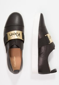Chaussures en cuir noir à enfiler avec une semelle blanche, dotées d'une bande métallique dorée portant l'inscription "Klein" et de détails élastiques sur les côtés.