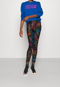 Blå sweatshirt med "VERSACE JEANS COUTURE" i rosa, kombinerat med svarta leggings som har färgglada, utsmyckade mönster och designdetaljer.