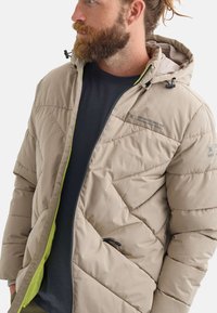 Chaqueta acolchada beige con capucha, que presenta un patrón de acolchado y un forro interior verde en contraste. Incluye cierre con cremallera y toggles ajustables.