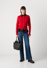 Red cardigan with buttons, layered over a black pinstriped blouse, paired with flared jeans and a black leather handbag. Platform shoes worn.