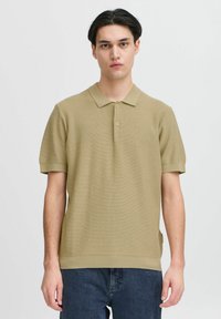 Khaki polo tričko s krátkým rukávem, rybkovaná textura a tři knoflíky. V kombinaci s tmavými džíny, úzký střih s bočním rozparkem.