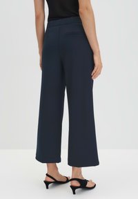 Pantalon large en tissu bleu marine foncé, doté d'une texture lisse et d'une poche au dos ; associé à des talons noirs à lanières.