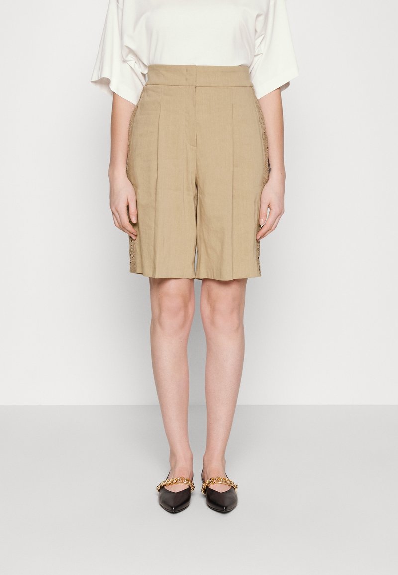 Beige op maat gemaakte shorts met plooien, zichtbare stoftextuur, schuin afgewerkte kantdetails. Gecombineerd met donkere, puntige schoenen met gouden kettingaccenten.