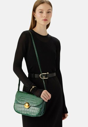 Grüne Handtasche aus geprägtem Leder im Krokodil-Look mit einer geschwungenen Klappe, goldenen Beschlägen und einem langen Schultergurt. Frau trägt ein schwarzes Top und einen Gürtel.