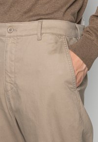 Pantaloni beige in misto cotone, con chiusura a pulsante, tasche frontali e una texture liscia. Dettagli di cucitura lungo le cuciture.
