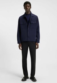 Giacca blu navy con due tasche sul petto, chiusura a bottoni e taglio dritto, abbinata a pantaloni neri e scarpe scure. Tessuto liscio.
