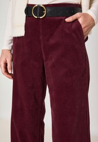 Pantaloni di velluto a coste bordeaux con un design a gamba larga, dotati di una cintura in pelle nera con una fibbia circolare dorata e tasche laterali.