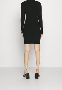 Robe en maille côtelée noire à manches longues, silhouette moulante et col rond, associée à des sandales noires à talons épais.