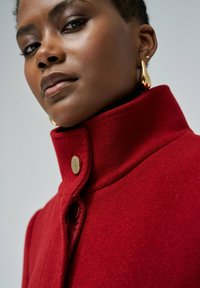 Crveni vuneni kaput s visokim ovratnikom, s zlatnim dugmetom za zatvaranje. Glatka tekstura i kroj koji ističe detalje majstorske izrade.