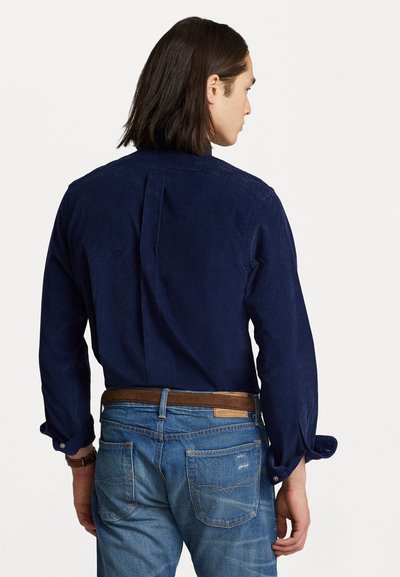 Mørkeblå cordfløyelskjorte med lange ermer, knapp mansjetter, ryggplym detalj. Kombinert med lyseblå denimjeans og brunt lærbelte.