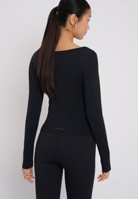 Schwarzes Langarmoberteil aus glattem Stoff mit quadratischem Ausschnitt und taillierter Passform, kombiniert mit gerippten schwarzen Leggings.