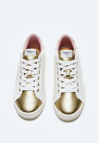 Sneakers bianche con accenti dorati metallici sulle punte e sui lati, caratterizzate da lacci bianchi, un'etichetta con il logo sulla linguetta e un rivestimento interno rosa.