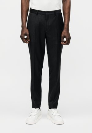 Mężczyzna w dopasowanych czarnych spodniach i białych trampkach stoi na jednolitym białym tle.