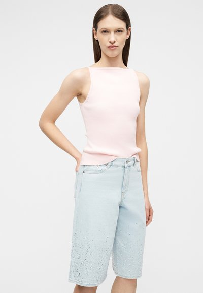 Νεαρή γυναίκα που φοράει αμάνικο ανοιχτό ροζ τοπ και ανοιχτό μπλε τζιν σορτς μέχρι το γόνατο με πουά λεπτομέρειες, στέκεται με το ένα χέρι στη μέση.