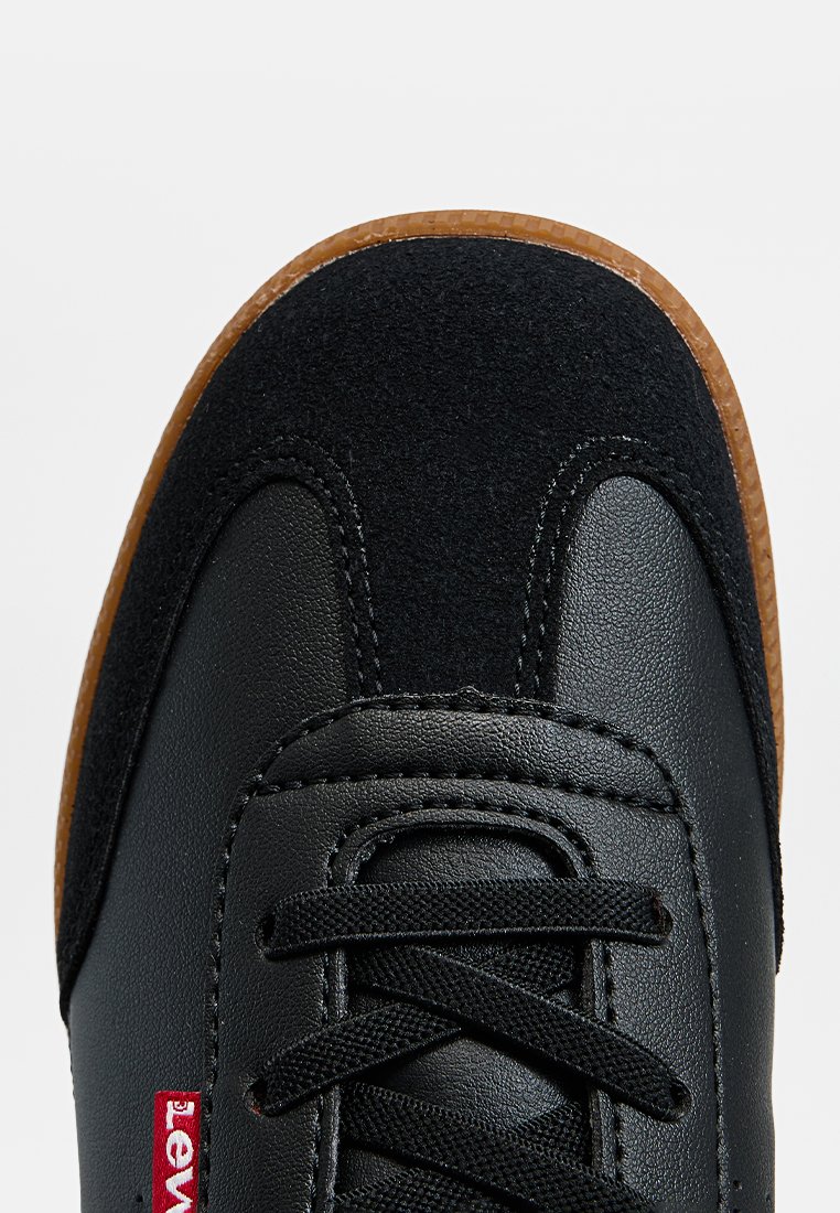 Zwarte sneaker met leren en vilt bovenwerk, ronde neus, zwarte veters en bruine rubberen zool. Heeft een rode merklabel aan de zijkant.
