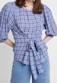 Lyse lilla ternet bluse med puffede ærmer, indviklet design og binding i taljen. Lavet af et struktureret stof, har den en rund halsudskæring.