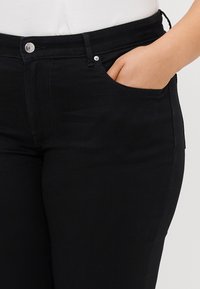 Fekete farmernadrág, elülső zsebbel, cipzárral és fém gombbal, valamint finom varrási részletekkel az illesztéseken és a derékon.