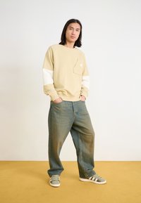 Felpa beige con maniche bianche, tasca frontale; abbinata a jeans blu larghi e sneaker grigio-verdi con strisce nere.