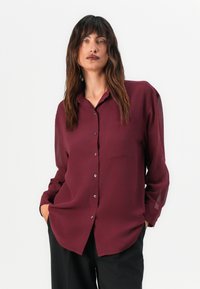 Camicia bordeaux con bottoni realizzata in tessuto leggero. Presenta un colletto a punta, lunghe maniche trasparenti e una tasca sul petto.
