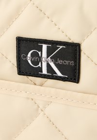 Tummanoranssi tikattu kangas, jossa musta suorakulmainen kangasmerkki valkoisella tekstillä: "CK" ja alla "Calvin Klein Jeans". Näkyvissä yksityiskohtainen ompelu.