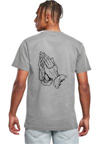 Siva bombažen T-shirt z črno ilustracijo zloženih rok z rožnim vencem, ki se nahaja na hrbtni strani. Kratke rokave, sproščen kroj.