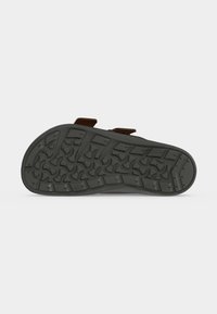 Μαύρη σόλα σανδαλιού με την κάτω όψη προς τα πάνω, που δείχνει πολύπλοκο σχέδιο πέλματος με γεωμετρικές και καμπυλωτές ραβδώσεις σε απλό γκρι φόντο.