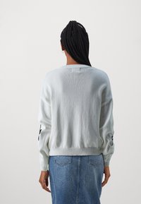Hellgrauer Strickpullover mit rundem Halsausschnitt, geripptem Saum und langen Ärmeln mit schwarzen Akzenten am linken Arm. Kombiniert mit einem Jeansrock.