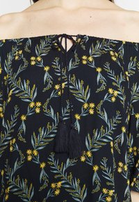 Top negro sin hombros con patrones florales en amarillo y verde, con un lazo de cuerda y borlas negras en el cuello.