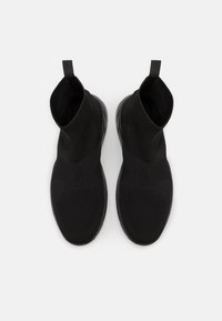 Pletené černé nazouvací boty s vysokým designem kotníku, strukturovanou látkou a hladkou gumovou podrážkou. Žádné viditelné logo ani ozdoby.