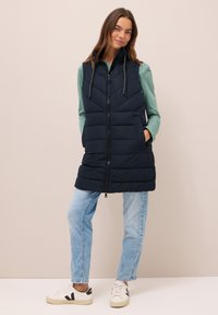 Gilet imbottito blu navy con zip frontale, colletto alto e due tasche laterali. Indossato sopra un top verde, abbinato a jeans azzurri chiari e sneakers.