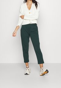 Weißer gerippter Cardigan mit V-Ausschnitt und einem einzelnen Knopf, kombiniert mit dunkelgrünen cropped Hosen und weißen Sneakern mit schwarzen Akzenten.