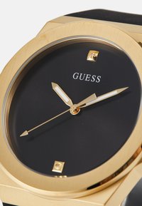 Reloj dorado con una esfera negra circular, marcadores de hora blancos y manecillas doradas. La caja es angular con un acabado texturizado y movimiento de cuarzo.