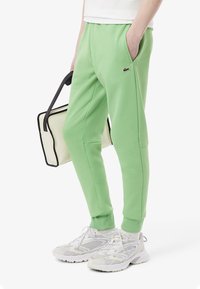 Grønne fleece sweatpants har en elastisk talje, sidelommer og et lille logo. De er parret med hvide sportsko og en kontrasterende taske.
