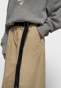 Un pull en maille côtelée gris associé à une jupe beige, ornée d'une ceinture noire à lanière et d'une broderie discrète "10" sur la jupe.