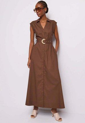 Vrouw die een mouwloze bruine maxi-jurk draagt met een brede riem, beige hakken zonder hiel en grote bruine zonnebril, staand tegen een witte muur.