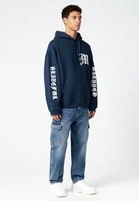 Marineblauwe hoodie met witte grafische tekst op de mouwen en borst, gecombineerd met lichtblauwe cargobroek en witte sneakers.