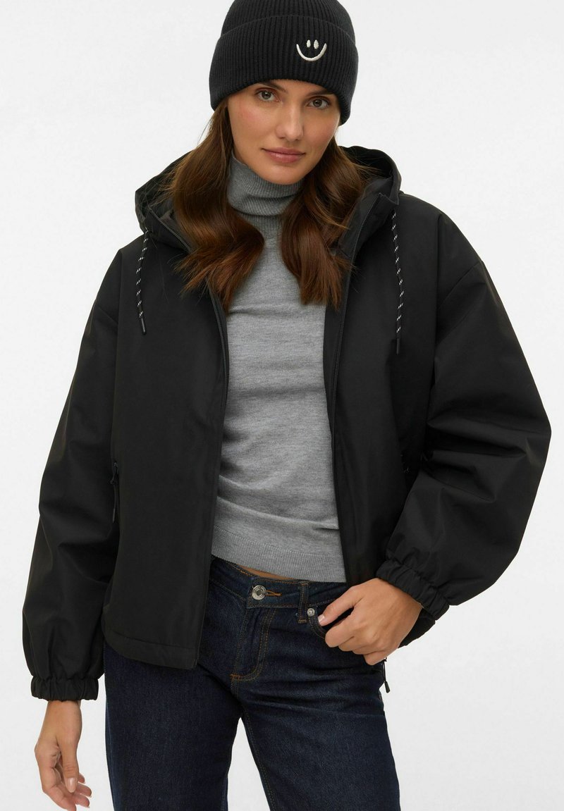 Kvinde iført en sort hue, sort hættetrøje med snore, grå højhalset sweater og mørkeblå jeans, stående foran en ensartet baggrund.