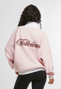 Felpa rosa con colletto e polsini bianchi. Testo grande "Felicious" in marrone sulla schiena. Morbida, vestibilità rilassata con spalle scese.