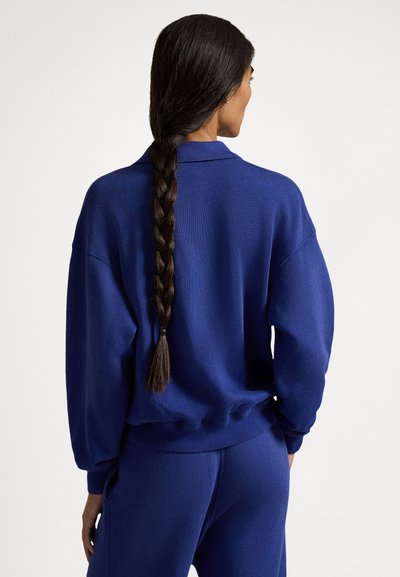 Temno modra prevelika majica z ovratnikom in rebrastimi manšetami, kombinirana z ujemajočimi temno modrimi hlačami. Gladek teksturiran in sproščen kroj.