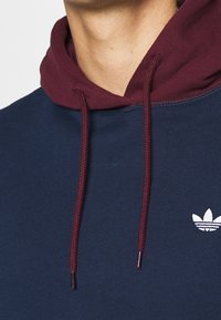 Mörkblå sweatshirt med en vinröd huva, med dragsko och en liten vit logotyp på bröstet. Mjuk textur och avslappnad design.