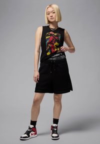 Camiseta sin mangas negra con gráfica colorida; pantalones cortos negros con cordón; zapatillas altas negras y rojas. Fondo gris liso.