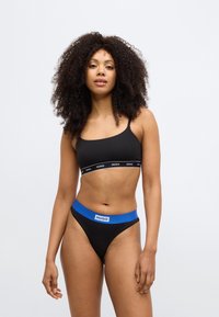 Must rinnahoidja valge ja musta "HUGO" logoga ribaga; sobivad mustad tangad sinise vöökohal oleva "HUGO" logoga. Sile tekstuur.