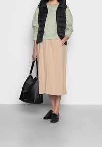 Gepolsterte schwarze Weste, hellgrünes Langarmoberteil, beiger Midikleid mit Taschen, schwarze Lederhandtasche und schwarze Slipper.