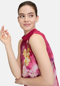 Ärmelloses Blumenoberteil mit hohem Kragen, lebhaftem pinken Stoff mit gelben und weißen Blumenmustern, weicher Textur und lockerem Schnitt.